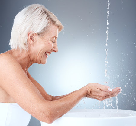Senior woman, water splash and cleaning hands in a basin for hygiene against a grey studio background with mockup space. Elderly model washing hand for, health, bacteria and clean body with liquidの写真素材