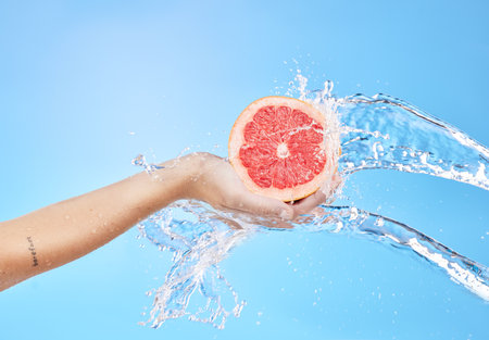 Hands, fruit and water splash for skincare wellness, cosmetics or nutrition against a blue studio background. Hand holding grapefruit for clean hygiene, hydration or vitamin C for body cleansingの写真素材