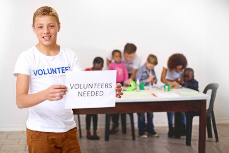 Help us help them. Portrait of a volunteer holding a volunteers needed sign with volunteers working with little children in the background.の写真素材