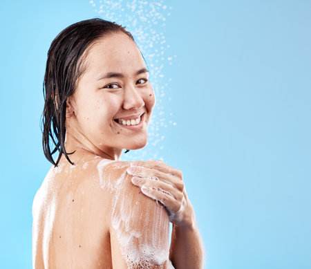 Shower, woman and hygiene for wellness, smile and happy with cleaning, water and soap against blue studio background. Portrait, young female and lady doing scrub, washing and health with happiness.の写真素材