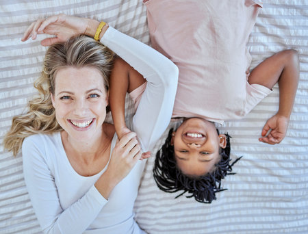 Top view, family and mother with foster girl on bed, relaxing and bonding. Love, care and adoption portrait of happy mom with black kid in bedroom, smiling and enjoying quality time together in houseの写真素材