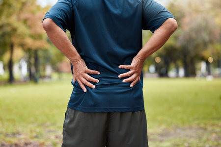 Exercise, fitness and back pain of man at park after workout. Break, healthcare or mature male runner with back injury, muscle pain and inflammation after exercising, running or training in natureの写真素材
