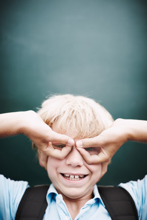Now you see me. A funny young boy making glasses with his hands against a classroom blackboard.の写真素材
