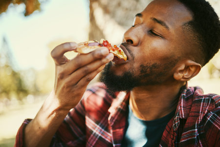 Fast food, hungry and black man eating pizza for delicious and yummy lunch break in park. Gen z, food and hunger of young person enjoying carbs pizza slice meal with satisfied face.の写真素材