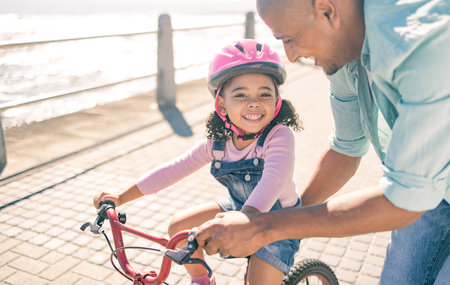 Black girl, learning and bike ride with dad at ocean promenade with smile, helmet or happy in sunshine. Kid, father and bicycle in training, safety or help for childhood development together outdoorの写真素材