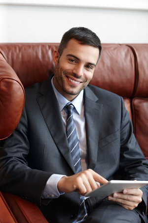 Hes a corporate success. A smiling young businessman sitting on a leather sofa while using a digital tablet.の写真素材