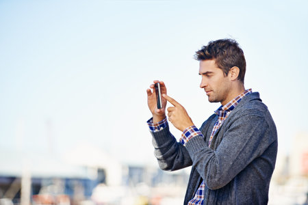 This ones a keeper. a handsome young man taking a photograph with his mobile phone phone outdoors.の写真素材