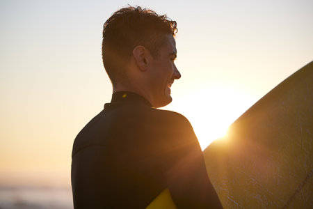 Fitness, sunset or man surfer with surfboard for exercise, workout or training wellness at the beach, ocean or sea with smile. Happy, freedom or lens flare male athlete for water sports motivationの写真素材