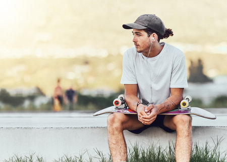 Young man, skateboard and relax outdoor, thinking or chill being casual, trendy and earphones. Mockup, male skater or idea for break, calm or cool look being edgy, content or hipster on weekend styleの写真素材