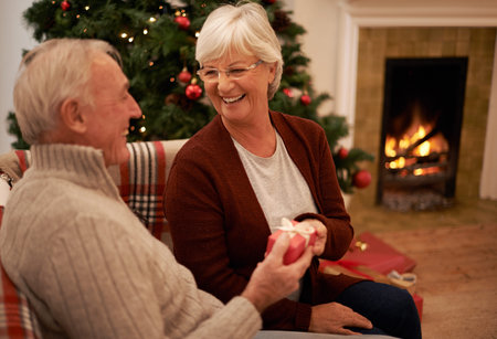 Christmas, gift and love with a senior couple in celebration while sitting in the living room of their home together. Tradition, present and bonding with a mature man and woman in the festive seasonの写真素材
