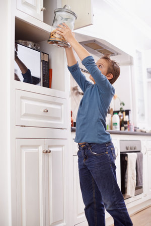 Mom will never know...A young boy reaching for a cookie jar in the top kitchen cupboard.の写真素材