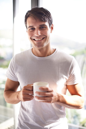 Theres nothing like a fresh cup of coffee in the morning. Portrait of a handsome young man drinking coffee while standing at his bedroom window.の写真素材