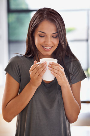 Starting the day with a fresh cup...an attractive young woman standing in her kitchen while drinking coffee.の写真素材