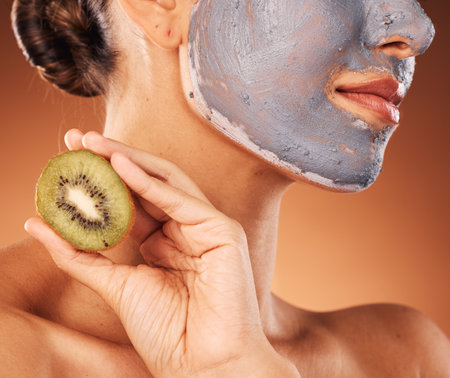Facial, mask and hand with kiwi for skincare of woman for wellness, beauty and self care routine. Cosmetic, spa and pamper lifestyle girl with natural treatment on brown studio background zoom.の写真素材