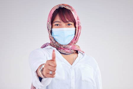 Covid face mask, muslim woman and thumbs up, portrait and vaccine yes vote, safety compliance and emoji, health risk and policy on studio background. Corona virus, thumb up and islamic female hijabの写真素材