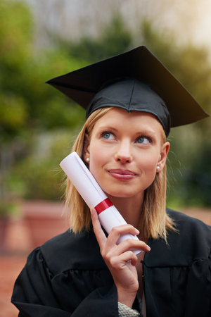 Go confidently in the direction of your dreams. a young college graduate looking thoughtful while holding her diploma.の写真素材