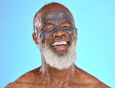 Face, water splash and senior black man in clay mask in studio isolated on a blue background. Cleaning, retired and elderly male from Nigeria washing off facial cosmetics for skincare or healthy skinの写真素材