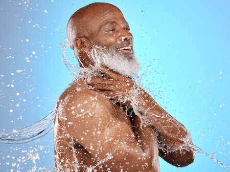 Beauty, water and cleaning splash of senior man for body care, hygiene and health lifestyle. Happy, shower and mature black model washing skin at studio blue background with satisfied smile.の写真素材