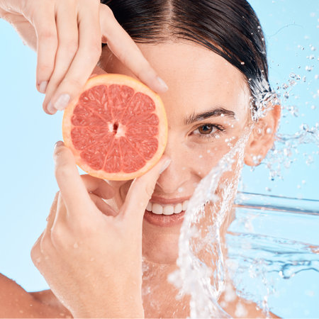 Face, water splash and woman with grapefruit, shower or skincare with happy smile, vitamin c or cleaning body. Female model, fruit or wellness in blue studio background with skin care or healthy skinの写真素材