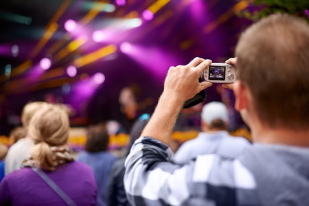 Living life at shutter speed. Rearview shot of a man in the crowd holding a camera to photograph a concert.の写真素材