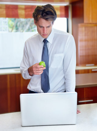 I only have time for a quick breakfast. A young man browsing on his laptop in the kitchen while enjoying a snack.の写真素材