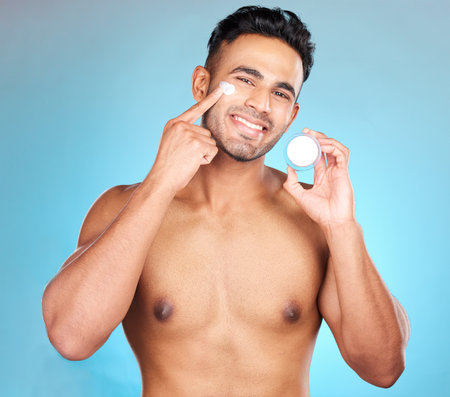Portrait, skincare and man in studio with face cream for wellness, pamper and cleaning on blue background. Face, beauty and facial, mask and product on guy model relax, luxury and grooming mockupの写真素材