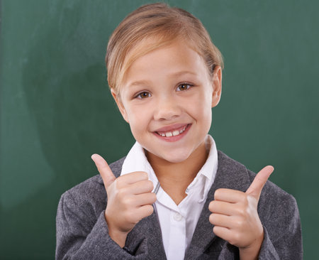 Shes ready to learn. Portrait of a young schoolgirl standing in front of a chalkboard and showing two thumbs up.の写真素材