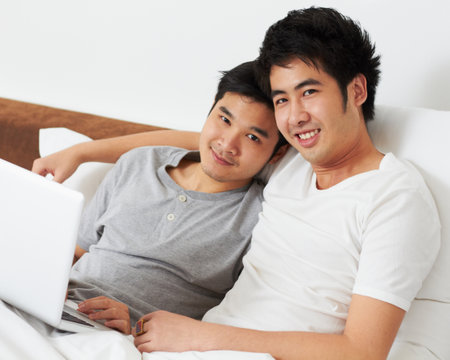 Just a day together at home. Portrait of a young gay couple relaxing with their laptop in bed.の写真素材