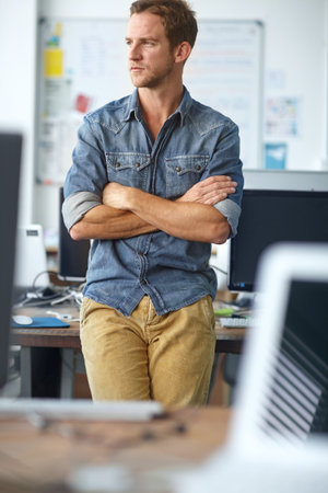 Thinking about his business decisions. A young casual businessman leaning against his desk in his...