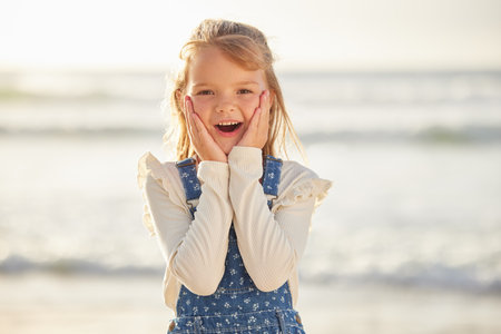 Close up view on a little young caucasian brunette girl making a shocked and astonished gesture with her hand while smiling and standing alone outside at the beach on a sunny summer day. Young girl lの写真素材