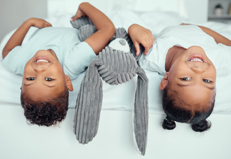 Adorable little sibling brother and sister lying upside down over the edge of a bed with a stuffed animal bunny. Cute little girl and boy having fun together at home in the morningの写真素材