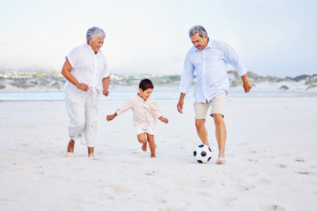 Active grandparents playing soccer on the beach with their grandson. Little boy kicking ball with his grandmother and grandfather on shoreの写真素材