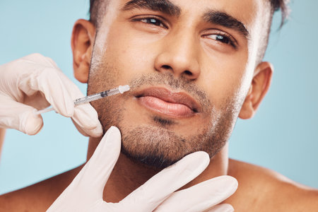 Handsome young indian man receiving a botox injection against a blue studio background. Mixed race guy getting treatment to reduce ageing and fill facial wrinklesの写真素材