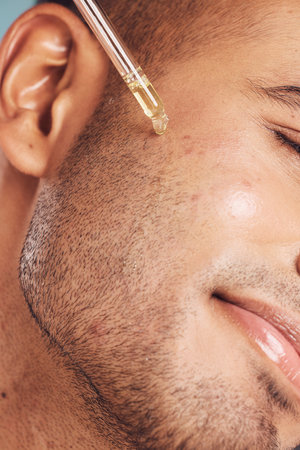 Closeup of one mixed race man using a dropper to apply serum oil to his skin and face against a blue studio background. Guy using a moisturising aftershave product for healthy, smooth and soft skinの写真素材