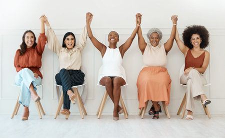 Group of five happy young businesswomen sitting against a wall in an office and holding hands in unity. Businesspeople smiling holding hands and sitting in a row together inside at workの写真素材