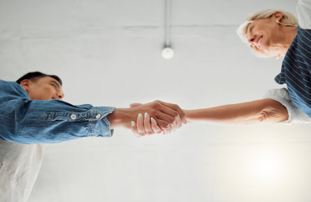 Two clothing designers shaking hands while working together at a shop. Mature caucasian woman and young asian man giving each other a handshake at work. Colleagues greeting at workの写真素材