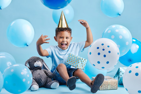 Excited little boy celebrating his birthday with balloons and gifts on a blue studio background. Cute kid wearing a party hat and sitting with presents and a stuffed bunnyの写真素材