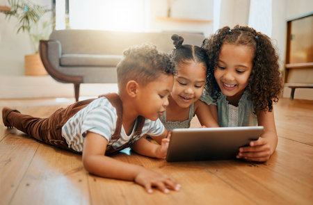 Three happy mixed race siblings relaxing on the lounge floor together while using a digital tablet. Three kids playing games on a digital device at homeの写真素材