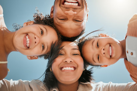 Portrait of a happy young mixed race family standing together in huddle smiling and looking down at camera on a sunny day. Faces of cheerful parents and two daughters embracing each other from belowの写真素材