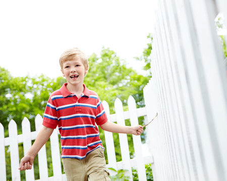 Energetic and excited in his backyard. A cute young boy running a stick along a white picket fence in his backyard.の写真素材