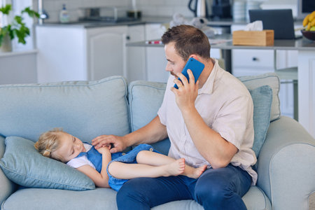 Sick little girl lying on sofa while dad uses cellphone to call doctor. Worried father on the phone to a healthcare professional while his ill daughter sleeps. Caring parent talking to family doctorの写真素材