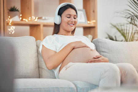 Pregnant, music and relax with a woman sitting on a sofa in their living room of her home expecting a baby. Pregnancy, wellness and stomach with a young female streaming audio alone in her apartmentの写真素材