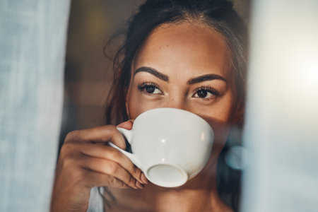 A beautiful young Hispanic woman enjoying a warm cup of coffee for breakfast. One mixed race female drinking tea while looking at the view from a window in her apartmentの写真素材