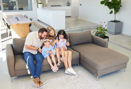 Playful caucasian parents tickling their daughters while relaxing together on a sofa at home. Carefree little girls spending time with loving parents. Joyful children while having fun with parentsの写真素材