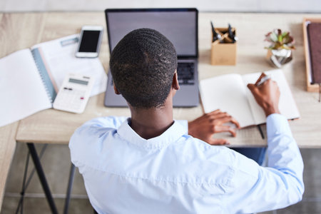 Top view, black man and writing with laptop, planning schedule and online reading in office. Nigerian male, entrepreneur and make notes for marketing strategy, search internet and ideas for business.の写真素材