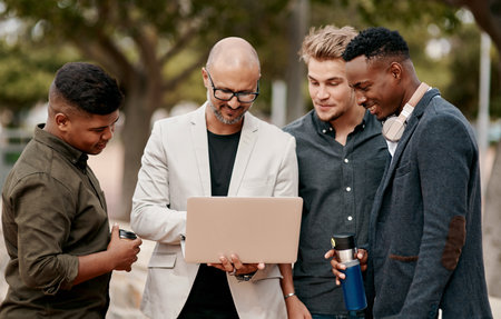 A team leader coaching his staff outdoors, standing and using a laptop. A group of male designers working on a start up project, looking at content on a computer outside, teamwork in the open air.の写真素材