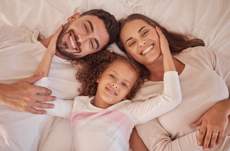 Portrait of happy love parents and daughter with smile in bed while relax in the morning. Family or mom, dad and child lying in bedroom having playful fun and bonding with happiness at home togetherの写真素材