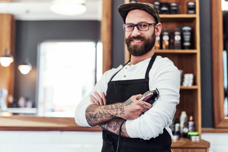 Stop by for the hippest haircuts in the city. Portrait of a handsome young barber posing with his arms folded inside his barbershop.の写真素材