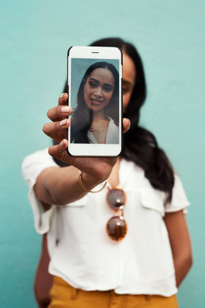 You cannot travel and not take a selfie. Cropped portrait of an attractive young woman standing and using her cellphone to take a selfie against a blue background.の写真素材