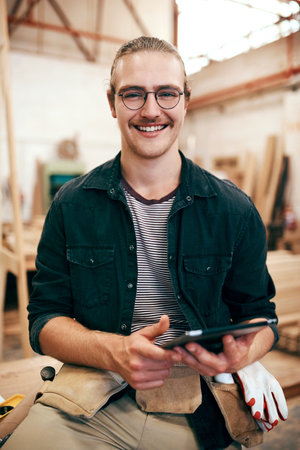 This device is my little assistant around here. Portrait of a handsome young carpenter using a digital tablet while working inside his workshop.の写真素材